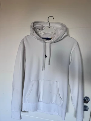 Vit hoodie från Polo Ralph Lauren - Säljer en stilren vit hoodie från Polo Ralph Lauren med klassisk huva och dragsnören. Den har en känguruficka framtill och den ikoniska Polo-loggan broderad på bröstet. Hoodien är gjord i mjukt bomullsmaterial och har ribbade muddar vid ärmar och nederkant. Passar för någon mellan 170-180