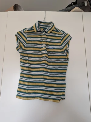 Randig pikétopp från BDG Urban Outfitters - Säljer en kortärmad pikétopp från BDG Urban Outfitters i grönt, gult, vitt och blått. Toppen har krage, knappar framtill och ett randigt mönster. Perfekt för dig som gillar retrovibbar och sköna material.