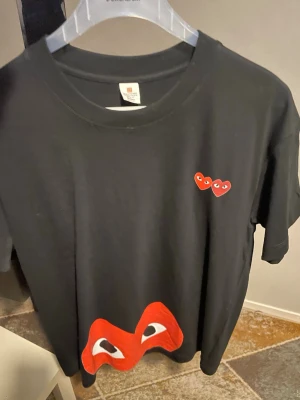 Svart t-shirt med röda hjärtan - Cool svart t-shirt med två små röda hjärtan med ögon på bröstet och ett stort rött hjärta med ögon längst ner. Perfekt för dig som gillar grafiska tryck och vill sticka ut lite extra.