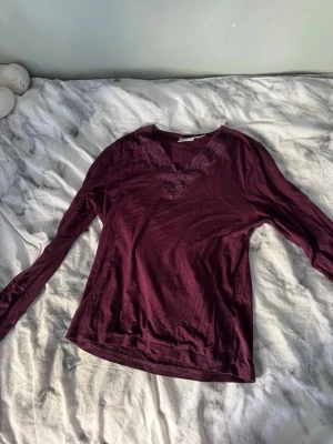 Vinröd långärmad topp med spets - Vinröd långärmad topp med v-ringning och dekorativ spetsdetalj vid halsen. Tillverkad i mjukt material som känns skönt mot huden. Perfekt för dig som gillar en enkel men ändå snygg stil.