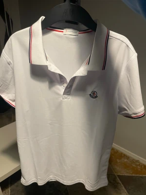 Vit Moncler pikétröja med ränder - Klassisk vit pikétröja från Moncler med korta ärmar och krage. Tröjan har röda och blå ränder på kragen och ärmsluten samt Moncler-logga på bröstet. Perfekt för en clean och sportig stil.