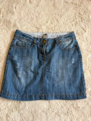 Low waist jeans kjol  - Snygg blå jeanskjol i klassisk modell med fem fickor och bälteshällor. Kjolen har en knapp och dragkedja framtill samt slitna detaljer för en trendig look. Perfekt till vardags och enkel att matcha med olika toppar.