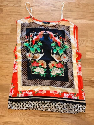 Färgglad linnetopp med fågelmönster - Säljer ett färgglatt linnetopp med ett stort, detaljerat mönster med tranor, blommor och blad i rött, grönt, vitt och svart. Toppen har en unik design med orientaliska inslag och kontrasterande bårder längst ner. Strl 36