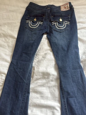 True religon joey broderade fickor - Snygga blå bootcut jeans med dekorativa broderier på bakfickorna och guldfärgade knappar. Jeansen har klassisk femficksmodell och är tillverkade i ett mjukt denimtyg som sitter skönt. Perfekta för dig som gillar en retroinspirerad look. Aldrig använda