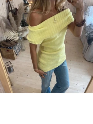 Ljusgul offshoulder stickad topp - Trendig ljusgul stickad topp med bred offshoulder-krage. Toppen är ärmlös och har en slim passform som framhäver figuren. Perfekt att styla med jeans för en cool och avslappnad look. Första bilderna är lånade