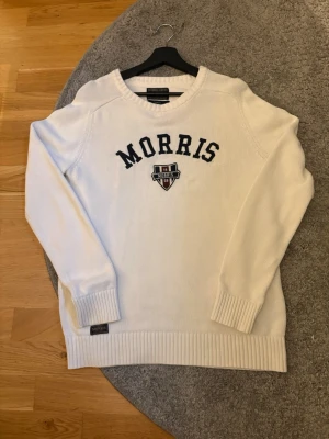 Vit stickad tröja från Morris - Snygg vit stickad tröja från Morris med mörkblå broderad logga och emblem på bröstet. Tröjan har rund halsringning, ribbade muddar och nederkant. Perfekt för dig som gillar klassisk college-stil.