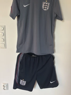 England Nike fotbollsträningsset - Säljer ett grått fotbollsträningsset från Nike med England-emblem på både tröja och shorts. Tröjan har korta ärmar och rund hals, shortsen har resår i midjan och diskreta röda detaljer längs sidorna. Perfekt för träning eller match!