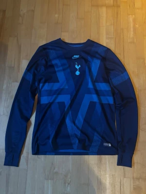 Marinblå långärmad Nike t-shirt - Marinblå långärmad t-shirt från Nike med Tottenham Hotspur-logga på bröstet. Tröjan har ett grafiskt mönster i olika blå nyanser och rund hals. Tillverkad i ett lätt och funktionellt material som passar perfekt till träning eller som supporterplagg.