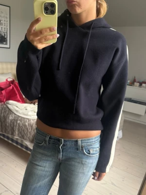 Mörkblå hoodie med vit rand - Säljer en mörkblå hoodie med huva och dragsnören. Hoodien har en vit rand längs hela ärmen och är croppad i modellen. Superfin