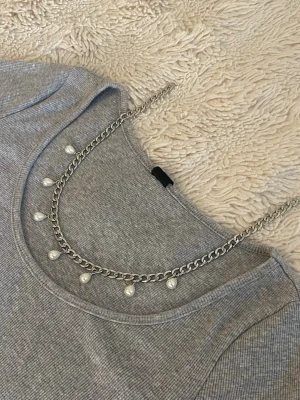 Silverfärgat halsband med pärlor - Snyggt halsband i silverfärgad rostfritt med kedja och vita pärlor som hänger längs hela framsidan. Perfekt för att ge en elegant touch till din outfit. Klassisk design som passar till många olika stilar.