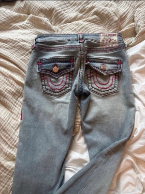 True Religion ljusblå jeans - Säljer ett par ljusblå jeans från True Religion med ikoniska bakfickor, rosa och mörkbruna sömmar💓 Jeansen de är i storlek 24 och är jättefina och oanvända💓
