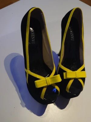 Svarta pumps med gula detaljer BINWENWU - Snygga svarta pumps från BINWENWU med gula lackdetaljer och rosett framtill. Skorna har peep-toe och hög klack, vilket ger en trendig och iögonfallande look. Perfekta för dig som vill sticka ut med färg och stil.