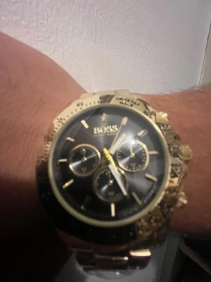 Guldfärgad klocka från BOSS - Stilren klocka från BOSS med guldfärgat armband och boett. Urtavlan är svart med guldiga detaljer och tre mindre subdials. Klockan har en klassisk rund form och metallarmband som ger en lyxig känsla.