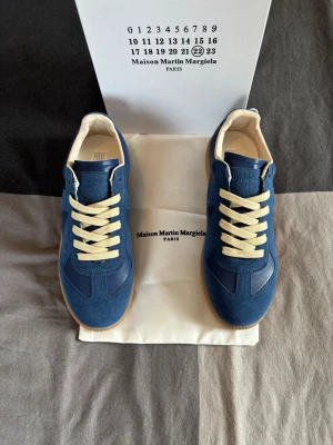 Blå Maison Margiela sneakers - Snygga blå sneakers från Maison Margiela med beigea skosnören och gummisula. Ovandelen är i mocka och läder, och insidan är fodrad i ljust material. Klassisk låg modell med stilren design och diskreta detaljer.