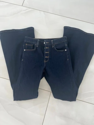 Mörkblå bootcut jeans med knappar - Säljer ett par mörkblå bootcut jeans med hög midja och snygga knappar framtill. Jeansen har klassiska fickor och är tillverkade i ett robust denimtyg. Superskönt material.