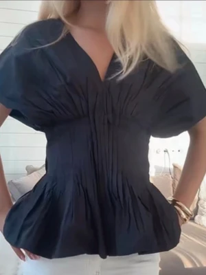Svart v-ringad blus med puffärm - Snygg svart blus med v-ringning och puffiga korta ärmar. Blusen har markerad midja och volang nedtill som ger en trendig siluett. Materialet ser ut att vara lätt och något glansigt, perfekt för att lyfta vilken outfit som helst. Helt slutsåld på nakd 