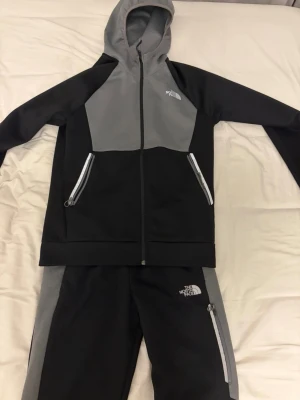 Svart & grå tracksuit från Nort Face - Snyggt loungewear-set från The North Face i svart och grått. Setet består av en hoodie med dragkedja och två fickor samt matchande byxor med dragkedjefickor. Perfekt för chill dagar hemma eller när du vill vara bekväm men ändå se fräsch ut.