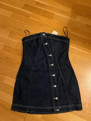 Mörkblå jeansklänning med knappar - Snygg mörkblå jeansklänning med dekorativa knappar framtill. Klänningen har figurnära passform och kontrastsömmar som ger en cool look. Perfekt för dig som gillar denim och vill ha en enkel men trendig stil.