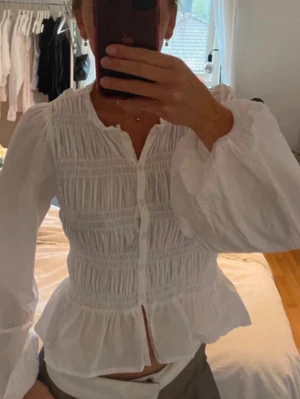 Vit blus med puffärm och smock - Säljer en vit blus med smockdetaljer och knappar framtill. Blusen har långa puffärmar och volang nedtill, vilket ger en romantisk och trendig look. Perfekt att styla med jeans eller kjol för en chic outfit.