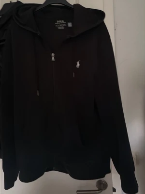 Svart hoodie från Polo Ralph Lauren - Svart hoodie från Polo Ralph Lauren med dragkedja framtill och klassisk logga broderad på bröstet. Hoodien har snörning i huvan och är tillverkad i mjuk bomull. Perfekt för en avslappnad och stilren look. 2000 nypris