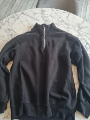 Svart halfzip sweatshirt - Svart sweatshirt med halv dragkedja och hög krage. Tröjan har ribbade muddar vid ärmslut och nederkant samt mjukt fleecefoder på insidan. Perfekt för en avslappnad och stilren look.
