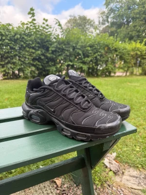 Nike Air Max Plus - Svarta Nike Air Max Plus sneakers med Tn-detalj på hälen. Skorna har en sportig design med vågiga linjer, mesh- och syntetmaterial samt kraftig sula med synliga luftkuddar. Perfekta för dig som gillar streetwear och vill ha en ikonisk look.