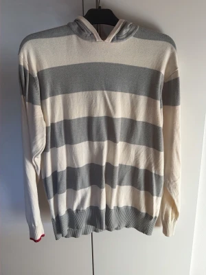 Randig hoodie i vitt och grått - Säljer en stilren hoodie med breda horisontella ränder i vitt och ljusgrått. Tröjan har huva och ribbade muddar vid ärmslut och nederkant. Perfekt för dig som gillar en avslappnad och enkel look. Materialet känns mjukt och lätt.