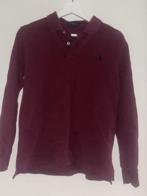 Vinröd långärmad skjorta  från Polo Ralph Lauren - Säljer en vinröd långärmad henley från Polo Ralph Lauren med klassisk krage och knappar vid halsen. Skjortan har en liten broderad logga på bröstet och är gjord i mjuk bomull. Perfekt för en avslappnad men snygg stil.