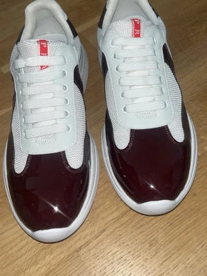 Prada sneakers i vinrött och vitt - Snygga sneakers från Prada med vinröd lackad tå och sidor, vita meshpartier och vita snören. Skorna har en chunky vit sula och röda Prada-detaljer på plösen. Perfekt för dig som vill sticka ut med en lyxig och modern look.