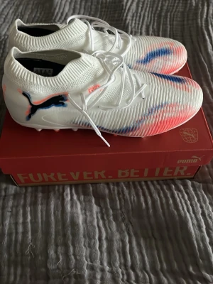 Puma Future 8 Match MG fotbollsskor - Säljer ett par vita Puma Future 8 Match MG fotbollsskor med svarta och röda detaljer samt blå inslag. Skorna har stickad ovandel för bra passform och snörning för stabilitet. Perfekta för spel på konstgräs och har en modern, sportig look. Dem är endast använda en gång pga köpte ett par andra. 