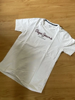 Vit Pepe Jeans t-shirt med tryck - Säljer en vit t-shirt från Pepe Jeans med mörkrött logotryck på bröstet och rund hals. Klassisk passform och korta ärmar. Perfekt till jeans eller shorts för en clean och enkel stil.