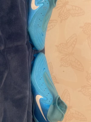 Blå Nike fotbollsskor med vit swoosh - Säljer ett par blå fotbollsskor från Nike med vit swoosh på sidan. Skorna har en strumpliknande öppning och mönstrad ovansida med små ventilationshål. Perfekta för dig som vill ha stilrena och moderna skor på planen. Skulle kunna sänka priset till 150 kr ! Acceptera också prisförslag!!!