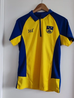 Blågul sporttröja från 361° 3XL - Liten i storlek!Snygg blå och gul sporttröja från 361° med kort dragkedja framtill och krage. Tröjan har korta ärmar och är tillverkad i ett lätt, ventilerande material. Perfekt för träning eller lagaktiviteter. Svensk landslagskänsla med tryckt emblem på bröstet.