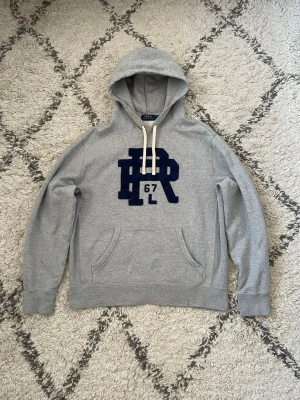 Ralph Lauren hoodie - Snygg grå hoodie med stor mörkblå RL 67-logga på bröstet och vita dragsnören i huvan. Klassisk känguruficka framtill och ribbade muddar vid ärmar och nederkant. Perfekt för en avslappnad och sportig stil. L sitter som M