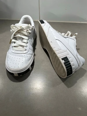 Vita Puma sneakers med svarta detaljer 38 - Snygga vita sneakers från Puma med svarta detaljer på sulan och perforerad tå. Skorna har klassisk låg profil, vita skosnören och är tillverkade i läder med gummisula. Perfekta för en streetwear-look och passar till det mesta.