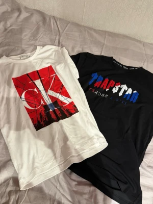 Två trendiga t-shirts med tryck - Säljer två snygga t-shirts, en vit Calvin Klein med stort rött och svart grafiskt tryck och en svart Trapstar med broderad text i rött, vitt och blått. Båda har rund hals och korta ärmar, perfekta för en cool streetwear-look.