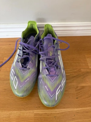 Lila och gröna F50 fotbollsskor - Säljer ett par snygga F50 fotbollsskor i lila och grönt med vita detaljer. Skorna har snörning och en låg profil, perfekt för snabba vändningar på planen. Insidan är limegrön och ovandelen har ett mönster med F50-tryck. Riktigt lätta och stilrena skor för dig som vill sticka ut.