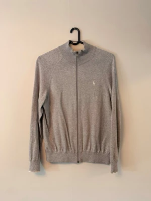 Grå ziptröja från Polo Ralph Lauren - Snygg grå ziptröja från Polo Ralph Lauren med hög krage och diskret logga på bröstet. Tröjan har långa ärmar, ribbade muddar och är tillverkad i mjukt material. Perfekt för lager på lager och enkel att matcha.