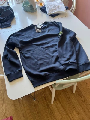 Mörkblå sweatshirt från Stone Island - Mörkblå sweatshirt från Stone Island med klassisk rund halsringning och ribbade muddar. Tröjan har den ikoniska Stone Island-loggan på vänster ärm och är tillverkad i mjuk bomull. Perfekt för en avslappnad och stilren look. Står XL men sitter som L