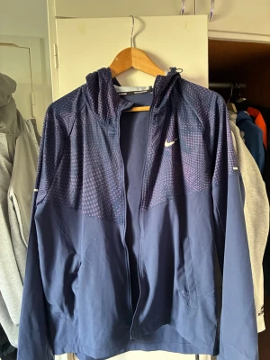 Mörkblå löparjacka från Nike - Snygg mörkblå löparjacka från Nike med hel dragkedja, huva och diskret mönster i lila och vitt på övre delen. Jackan har två sidofickor och Nike-logga på bröstet. Perfekt för löpning eller träning utomhus.