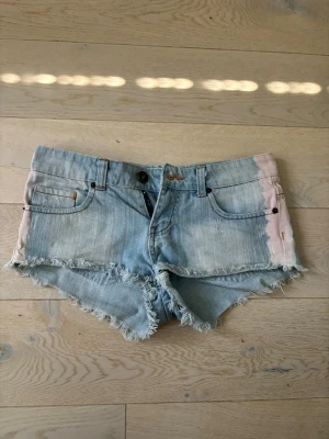 Ljusblå jeansshorts med rosa detaljer - Säljer ett par ljusblå jeansshorts med råa, fransiga kanter och rosa detaljer längs sidorna. Dragskedjan är sönder men knappen funkar! Dom är low rise i storlek XS, vet ej vart dom är ifrån.