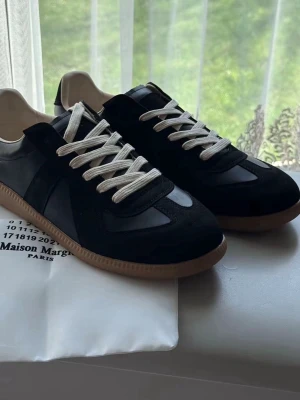 Maison Margiela svarta sneakers - Svarta sneakers från Maison Margiela med beige sula och vita skosnören. Skorna har en mix av mocka och läderdetaljer, klassisk låg profil och gummisula. Perfekta för dig som gillar stilrena och tidlösa sneakers.