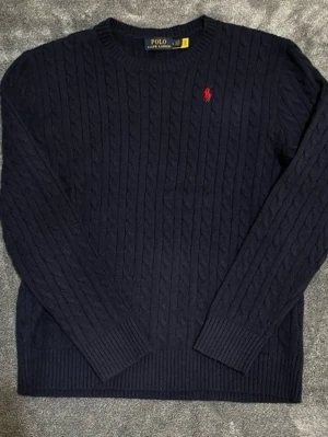 Mörkblå kabelstickad tröja Polo Ralph Lauren - Kabelstickad mörkblå tröja från Polo Ralph Lauren med rund halsringning och röd broderad logga på bröstet. Tröjan har långa ärmar och ribbade muddar vid ärmslut och nederkant. Perfekt för en klassisk och stilren look.