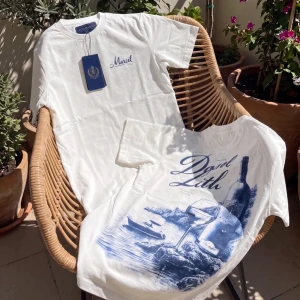 Vit t-shirt med blått tryck - Vit t-shirt med blått tryck framtill och stort konstnärligt motiv på ryggen med vinflaska, glas och havsmotiv. Rund hals och korta ärmar. Märket 'Martel' syns på bröstet. Perfekt för dig som gillar stilrena men ändå unika plagg.