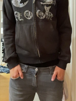 Aim for the moon hoodie (full zip) - Säljer en svart hoodie med dragkedja och huva. På framsidan finns stora tryck i strass med text och motiv, bland annat en tecknad figur och en måne. Hoodien har fickor framtill och ribbade muddar vid ärmar och nederkant. Perfekt för dig som gillar streetwear och detaljer.