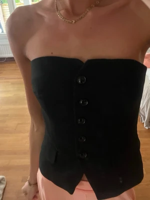 Svart korsett-topp med knappar - Snygg svart korsett-topp med knappar framtill och figurnära passform. Toppen är axelbandslös och har en stilren, elegant look med små fickdetaljer framtill. Perfekt att styla med både jeans och kjol för en trendig outfit.