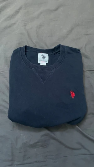 Mörkblå sweatshirt från U.S. Polo Assn. - Mörkblå sweatshirt med rund hals från U.S. Polo Assn. Tröjan har ett rött broderat logomärke på bröstet och ribbade muddar vid ärmslut och nederkant. Perfekt för en avslappnad och stilren look.
