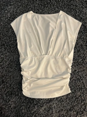 Vit v-ringad topp med rynkade detaljer - Säljer en vit topp med v-ringning och rynkade detaljer i sidan. Toppen är ärmlös och har en figurnära passform med snygg drapering. Perfekt att styla med jeans eller kjol för en trendig look.