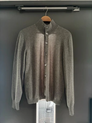 Brunello Cucinelli cardigan - Stilren grå kofta med ståkrage och knäppning framtill. Tillverkad i mjukt, tunt stickat kashmir och har ribbade muddar vid ärmslut och nederkant. Perfekt för lager på lager och enkel att matcha med olika outfits.