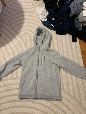 Grå hoodie från Sail Racing - Snygg grå hoodie från Sail Racing med dragkedja framtill och en vertikal bröstficka med logga. Hoodien har huva med justerbara snören och är tillverkad i mjukt material. Perfekt för chill dagar eller när du vill ha en clean look.